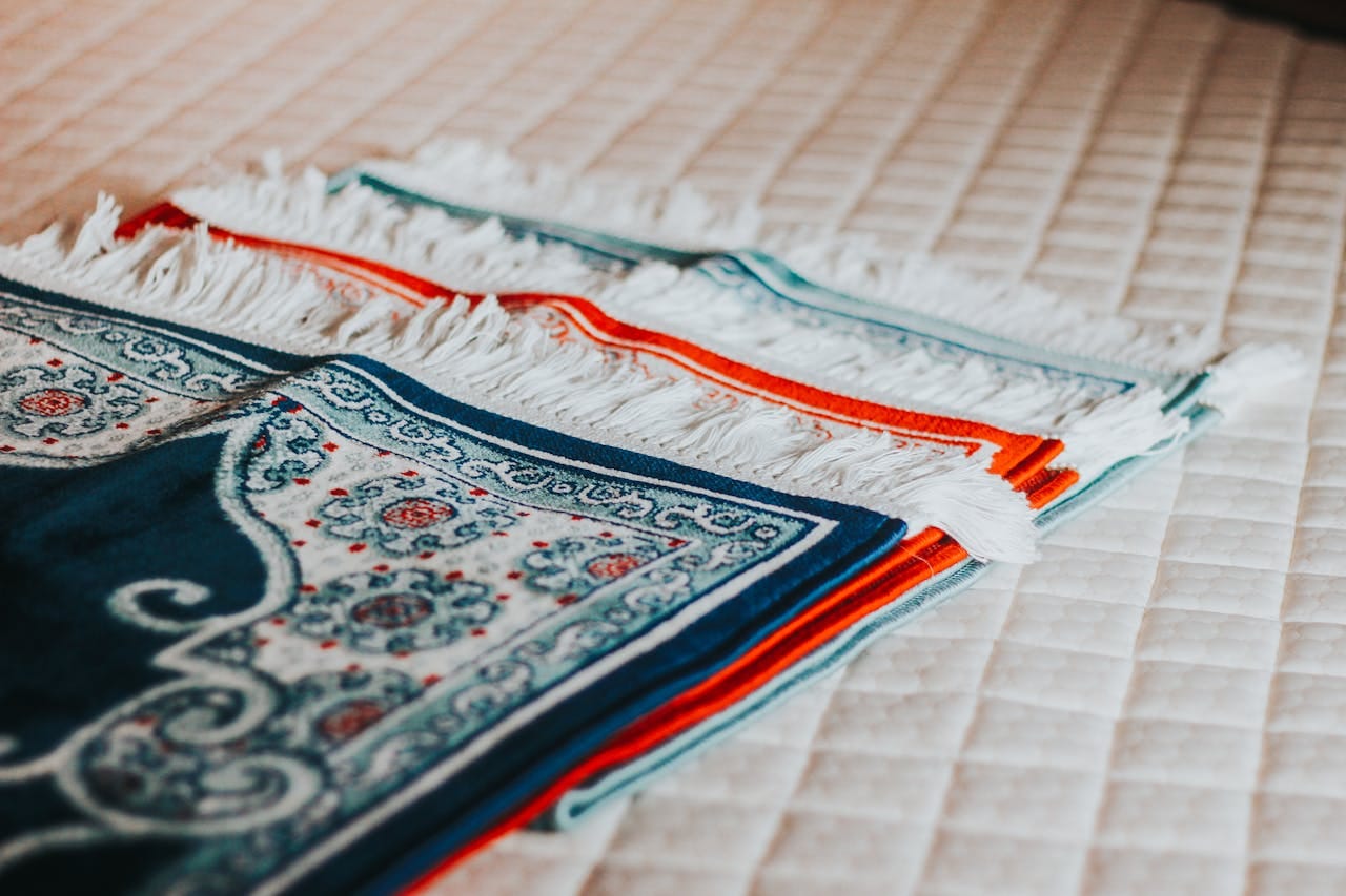 A close-up of vibrant, intricately designed prayer rugs with fringe detail laid on a quilted surface.